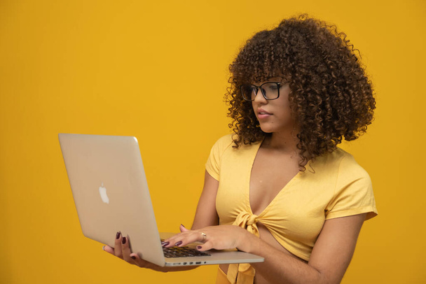 Young latin curly woman posing with laptop on bright yellow background - Фото, изображение