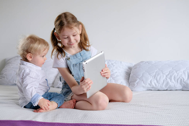 two children at home in bed are playing businessmen. A little girl is playing on a tablet with her little brother. - Foto, Imagem