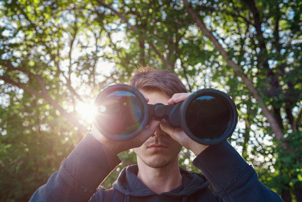 Explorer hiker young man in plaid shirt looking through binoculars on background of summer forest and sea bay outdoor, front view. Travel concept. - Fotografie, Obrázek