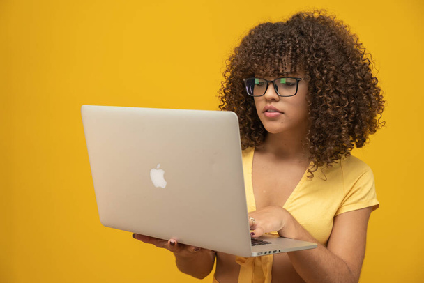 Молодая латинская кудрявая женщина позирует с ноутбуком на ярко-желтом фоне
 - Фото, изображение