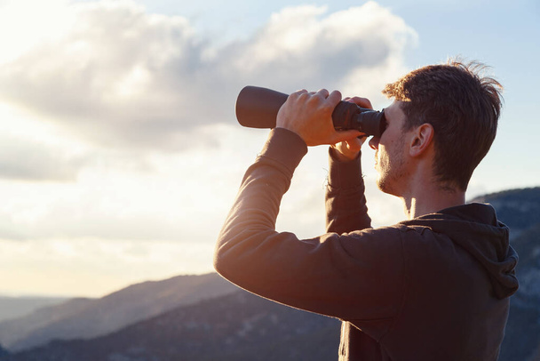 Mladý muž v horách se dívá dalekohledem. Nápis s kopírovacím prostorem. myšlenka a koncepce cestování a objevů, dobrodružství a svobody - Fotografie, Obrázek