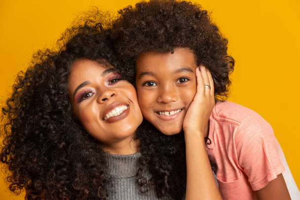 幼い息子を持つ若いアフリカ系アメリカ人の母親の肖像画。黄色の背景。ブラジルの家族. - 写真・画像