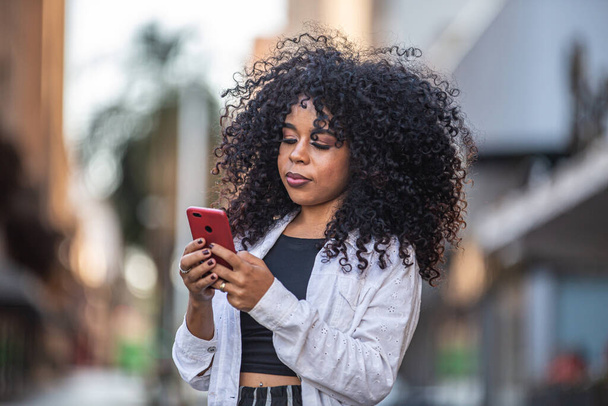 Νέοι σγουρά μαλλιά μαύρη γυναίκα περπάτημα χρησιμοποιώντας το κινητό τηλέφωνο. Στέλνω μηνύματα στο δρόμο. Μεγάλη πόλη. - Φωτογραφία, εικόνα