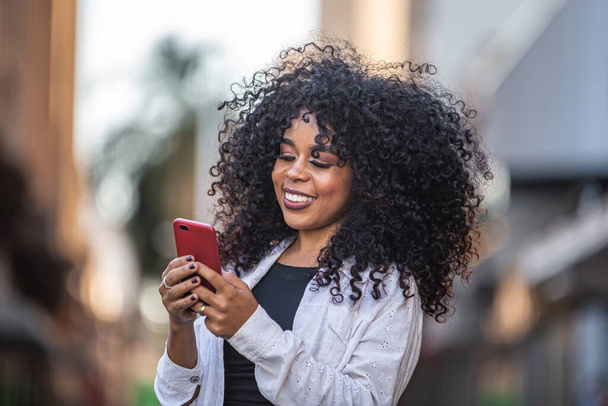 Jovem cabelo encaracolado mulher negra andando usando telefone celular. A enviar mensagens na rua. Cidade grande
. - Foto, Imagem