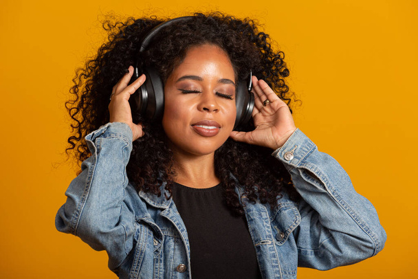 People, music, emotions concept. Delighted carefree female with Afro hairstyle dances in rhythm of melody, closes eyes listens loud song in headphones. Beautiful afro woman with her headphones - Foto, Imagem