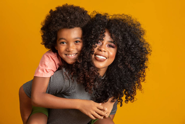 Portret van jonge Afro-Amerikaanse moeder met peuter zoon. Gele achtergrond. Braziliaanse familie. - Foto, afbeelding