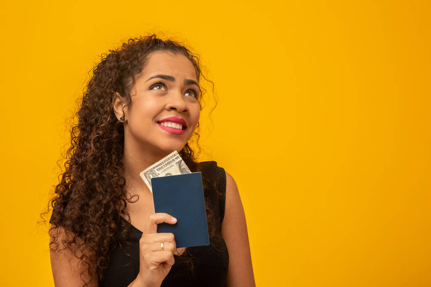 Belle jeune femme aux cheveux bouclés tenant un passeport et de l'argent, en pensant à son prochain voyage. Sur fond jaune
 - Photo, image