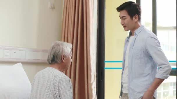 νεαρός ενήλικος ασιάτης γιος που μιλάει και παρηγορεί τον πατέρα του σε νοσοκομείο ή γηροκομείο - Πλάνα, βίντεο