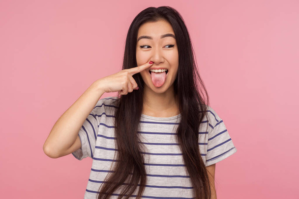 Portrait of funny comical girl with long hair in striped t-shirt picking nose, pulling boogers and showing tongue with humorous silly expression. indoor studio shot isolated on pink background - Photo, Image