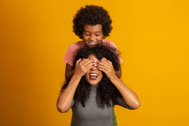 African American boy holding his mother closed eyes. Yellow background. Mothers Day. Brazilian family. - Фото, зображення