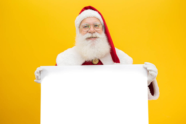 Happy Santa Claus looking out from behind the blank sign isolated on yellow background with copy space - Photo, Image