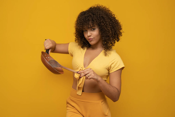 Молодая латинская кудрявая женщина позирует с сковородкой и лопаткой на ярко-желтом фоне
 - Фото, изображение