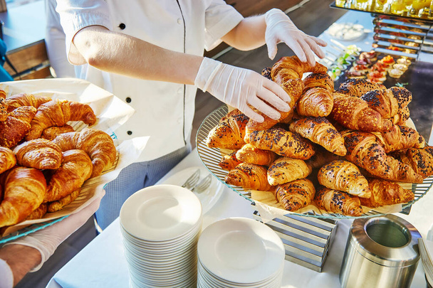  Hands in white gloves of two caters, one of which spreads croissants on a dish, and the other holds a dish with croissants - Photo, image