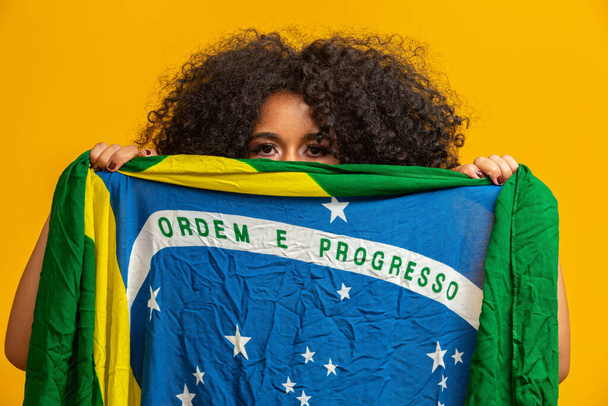Misterioso abanico de mujer negra sosteniendo una bandera brasileña en tu cara. Brasil colores en el fondo, verde, azul y amarillo. Elecciones, fútbol o política
. - Foto, Imagen