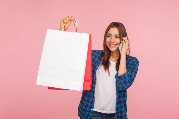 Portrait of glad smiling girl in checkered shirt holding shopping bags and talking on cellphone, making purchase, ordering delivery using mobile phone. indoor studio shot isolated on pink background - Foto, immagini