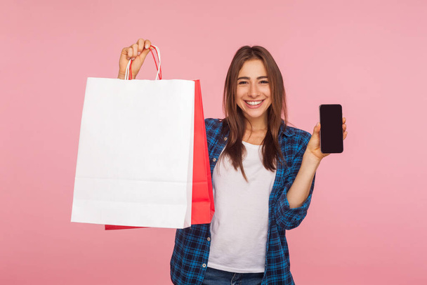 Portrait of happy smiling girl in checkered shirt holding cell phone and shopping bags, advertising online store on mobile device, good delivery application. studio shot isolated on pink background - Фото, изображение
