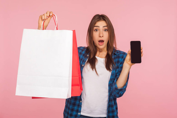 Portrait of surprised girl in checkered shirt holding cellphone and shopping bags, shocked by online store on mobile device, good delivery application. indoor studio shot isolated on pink background - Foto, imagen