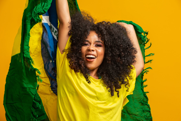Afro niña animando a la selección brasileña favorita, sosteniendo la bandera nacional en el fondo amarillo
. - Foto, Imagen