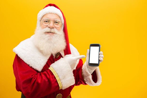 Άγιος Βασίλης με κινητό σε κίτρινο φόντο. Έννοια της επικοινωνίας, τηλεφωνικές εταιρείες και προωθητικές ενέργειες. - Φωτογραφία, εικόνα