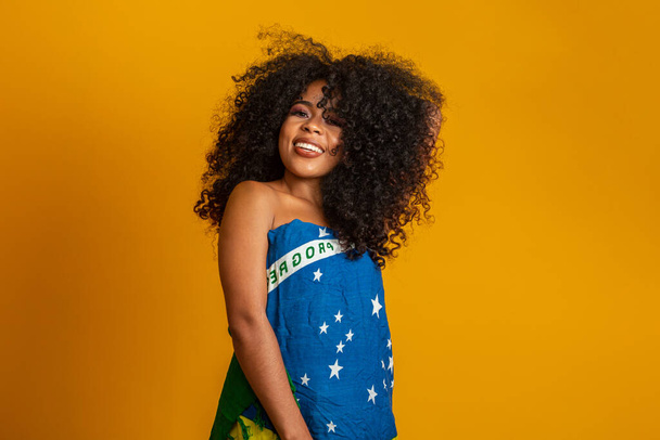 Happy black woman fan holding a Brazilian flag. Brazil colors in background, green, blue and yellow. Elections, soccer or politics. - Photo, Image
