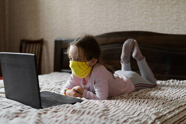 Concepto de aprendizaje a distancia en línea durante pandemia y cuarentena. Una niña se encuentra en casa en el sofá y mira las lecciones en línea
. - Foto, Imagen