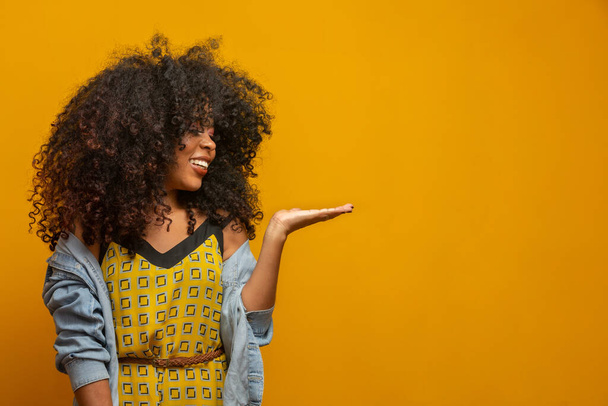 Cheerful Afro woman points away on copy space, discusses amazing promo, gives way or direction, wears yellow warm sweater, has pleasant smile, feels optimistic, isolated over yellow background. - Photo, Image