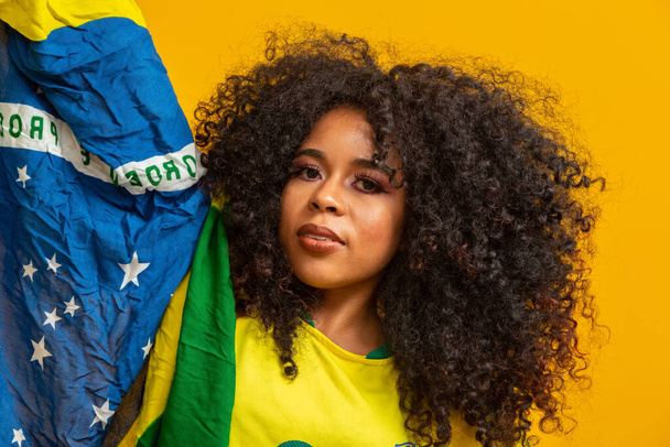 アフロの女の子は好きなブラジルのチームのために応援し、黄色の背景に国旗を保持. - 写真・画像