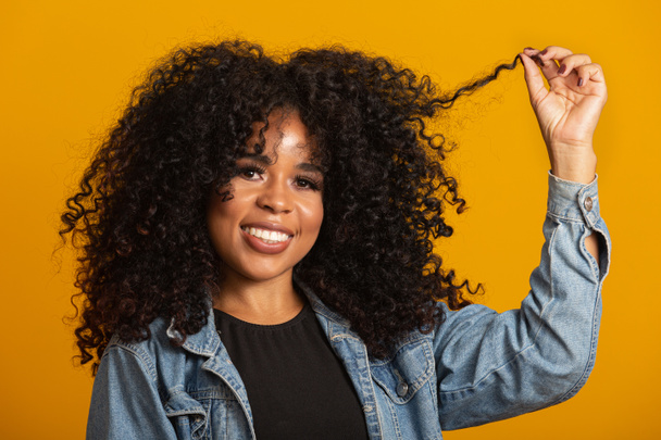Νεαρή Αφρο-Αμερικανίδα με σγουρά μαλλιά που κοιτάει την κάμερα και χαμογελάει. Χαριτωμένο afro κορίτσι με σγουρά μαλλιά χαμογελώντας κοιτάζοντας κάμερα. - Φωτογραφία, εικόνα