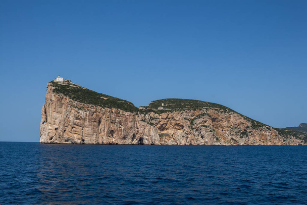 Calm blue water of Mediterranean sea, surrounding peninsula Capo Caccia with a white building on the top of the cliff. Bright blue sky. Sardinia, Italy. - Photo, Image