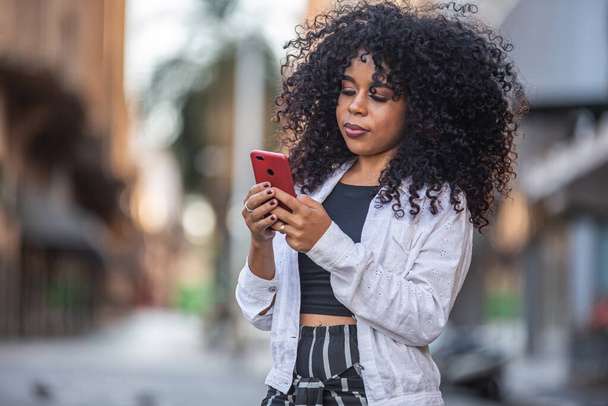 Jonge krullend haar zwarte vrouw loopt met behulp van mobiele telefoon. Ik sms op straat. Grote stad. - Foto, afbeelding