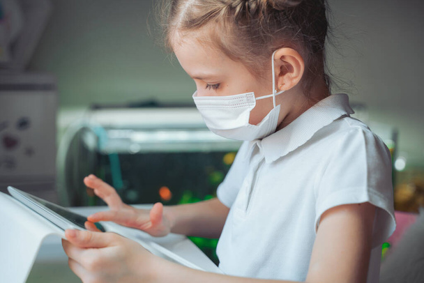 Caucasian preteen girl with medical mask on her face concentrated on her task with tablet. Concept of distance learning in isolation while coronavirus - Foto, afbeelding