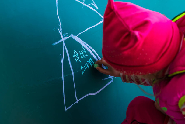 ピンクのジャケットの女の子が学校の黒板にチョークで家を描きます。彼女の手にはチョーク. - 写真・画像