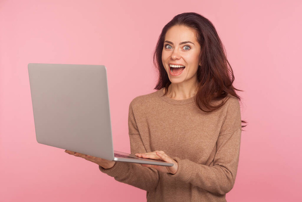 Portrait of young woman holding laptop and looking at camera with shocked expression, pleasantly surprised by computer software, shopping online. indoor studio shot isolated on pink background - Φωτογραφία, εικόνα