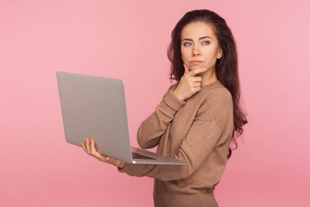 Inspiration, business idea. Portrait of young woman in casual sweater holding laptop and thinking over startup strategy with serious doubtful expression. indoor studio shot isolated on pink background - Foto, immagini