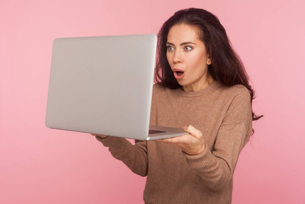 Portrait of amazed young woman standing with mouth open in surprise and reading shocking information on laptop, worried about mistake, software error. indoor studio shot isolated on pink background - Foto, Imagem