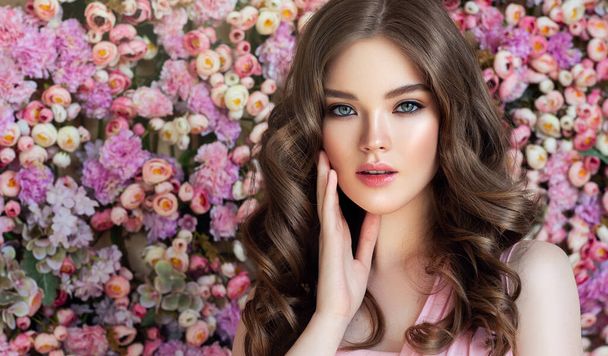 Brunette spring girl with long and shiny wavy hair . Beautiful model woman with curly hairstyle , background wall of flowers . Skin Care, beauty and spa - Fotoğraf, Görsel