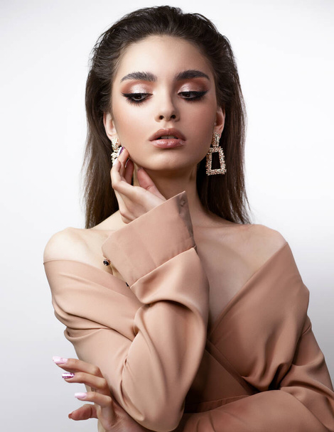 A woman in an elegant beige dress, evening make-up, holds her hand near her face. There are large earrings in the ears. She is looking down. - Foto, immagini