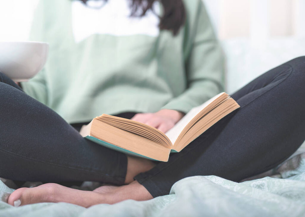 Jonge donkerharige vrouw zit in bed en leest haar lievelingsboek, drinkt koffie uit witte beker - Foto, afbeelding