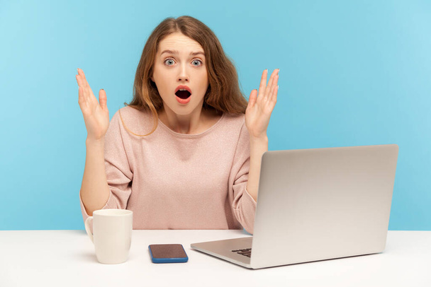 Oh my god, wow! Young woman with shocked expression looking at camera with open mouth and raising hands in surprise, working on laptop from home office. indoor studio shot isolated, blue background - Foto, immagini