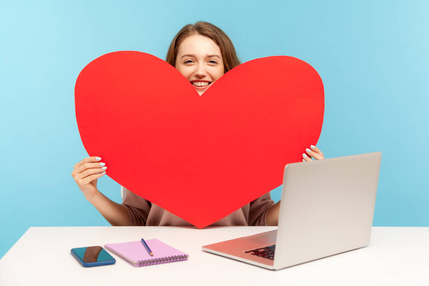 Online dating service. Beautiful woman holding big heart love symbol and smiling, sitting at desk with laptop, advertising website for romantic date. indoor studio shot isolated on blue background - Fotó, kép