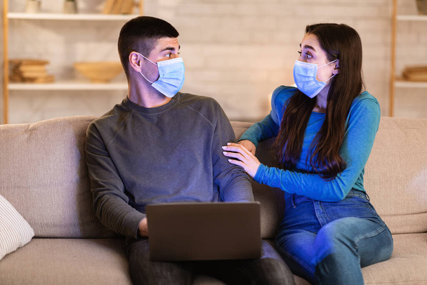Scared couple in face masks using laptop sitting in sofa - Photo, Image