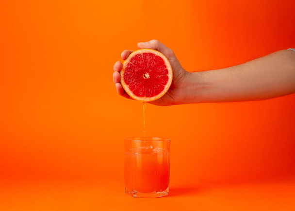 Detailně oříznutá fotografie ženy držící v rukou polovinu čerstvého zralého grapefruitu, skleněný pohár izolovaný na oranžovém pozadí. Lidé živý životní styl relaxovat dovolenou koncept. Zahájit kopírovací prostor - Fotografie, Obrázek