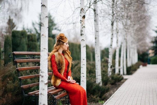 joven con el pelo rojo en un vestido rojo brillante en un banco en un parque vacío entre los abedules
 - Foto, imagen