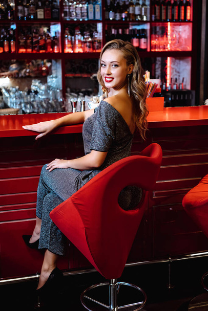 Kaunis tyttö loistavassa mekossa istuu baarissa. Nuori tyttö malli istuu punaisella tuolilla taustalla bar sisustus
 - Valokuva, kuva