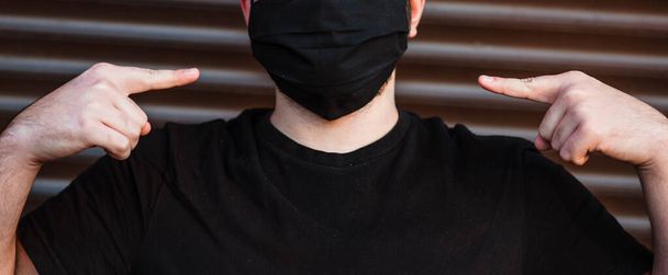 黒いTシャツとマスクの若い男が暗い壁の背景に隔離された。健康的なライフスタイル,病気の患者を治療,寒い季節の概念.彼らはコピースペースをモックする。滅菌面マスクを着用する. - 写真・画像