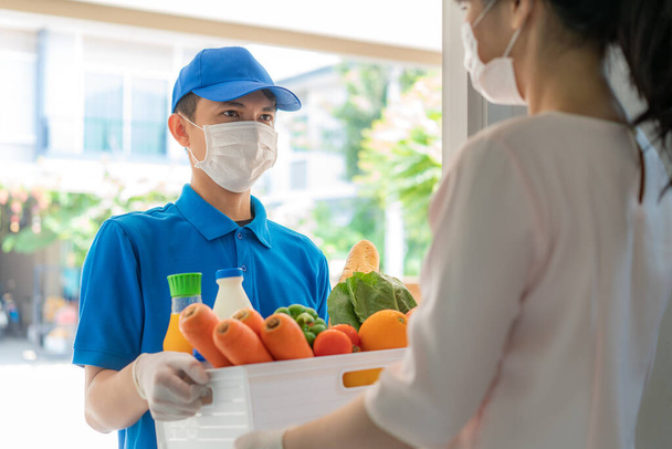 Azjatycki dostawca noszenia maski twarzy i rękawiczki z pudełka żywności, owoców, warzyw i napojów dać kobiecie kostiumy przed domem w czasie izolacji domu. - Zdjęcie, obraz