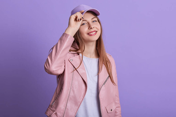 Closeup portrait of attractive woman wearing pink leather jacket, shirt and cap, lady touches her headdress and looking at camera with charming smile, posing isolated over lilac studio background. - Foto, Bild