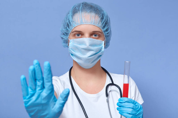 Picture of serious hard working doctor holding tube with blood for coronavirus test, making gesture, raising hand, wearing protective mask, hat and gloves, controling situation. Covid19 concept. - Fotó, kép