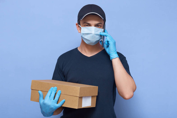 Horizontal picture of busy active delivery man looking directly at camera, wearing protective mask and gloves, holding box, working at online delivery services, talking over phone. Covid19 concept. - Photo, image