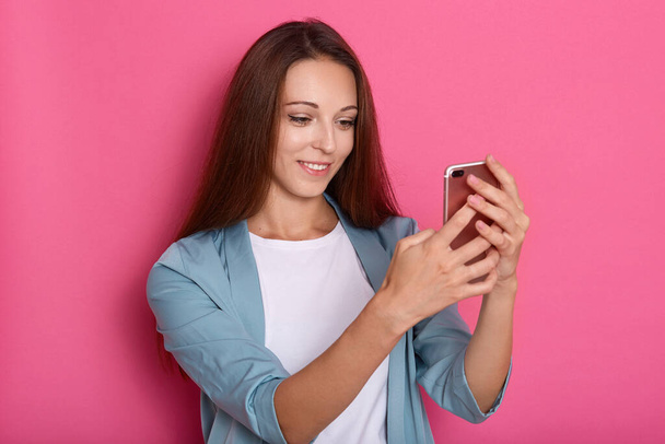Indoor picture of delighted sweet lovely young lady holding smartphone in both hands, making selfie, making videocall, talking over phone, having pleasant facial expression. Technology concept. - Fotoğraf, Görsel